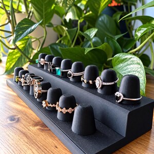 May include: A black tiered ring display stand with multiple cone-shaped holders, showcasing a variety of metal rings with diverse gemstone settings, including emerald-cut green stones, clear diamonds, and black accents arranged in rows.