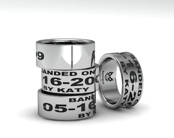 May include: A stack of four silver rings with black text. The rings are engraved with dates and the words "BANDED ON" and "BY KATY". One ring stands apart, displaying the "duckband" logo.