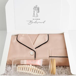 May include: A gift box containing a blush pink pajama set with black trim, a clear jar with a pearl hair clip and lip balm, and a card that reads "Will You Be My Bridesmaid" with a line drawing of a couple.