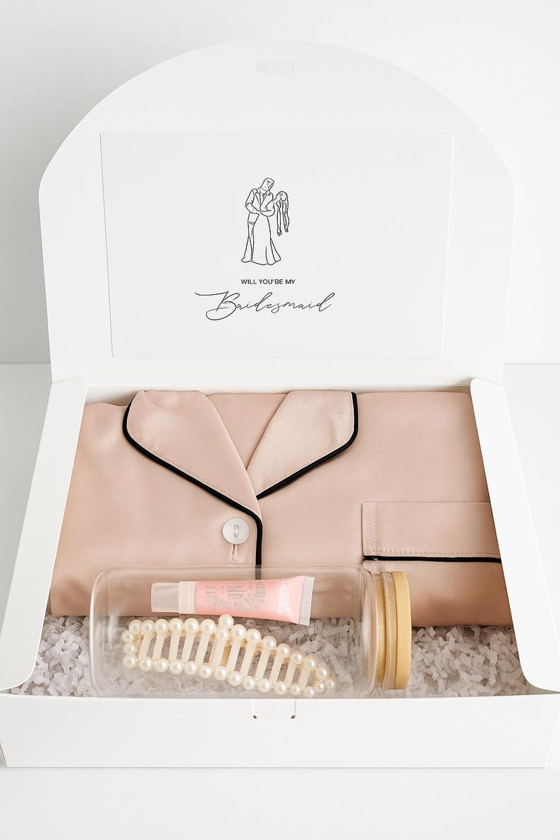May include: A gift box containing a blush pink pajama set with black trim, a clear jar with a pearl hair clip and lip balm, and a card that reads "Will You Be My Bridesmaid" with a line drawing of a couple.