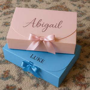 May include: Two gift boxes, one pink and one blue, stacked on top of each other. The pink box has the name "Abigail" in script, and a pink bow. The blue box has the name "LUKE" and a blue bow.