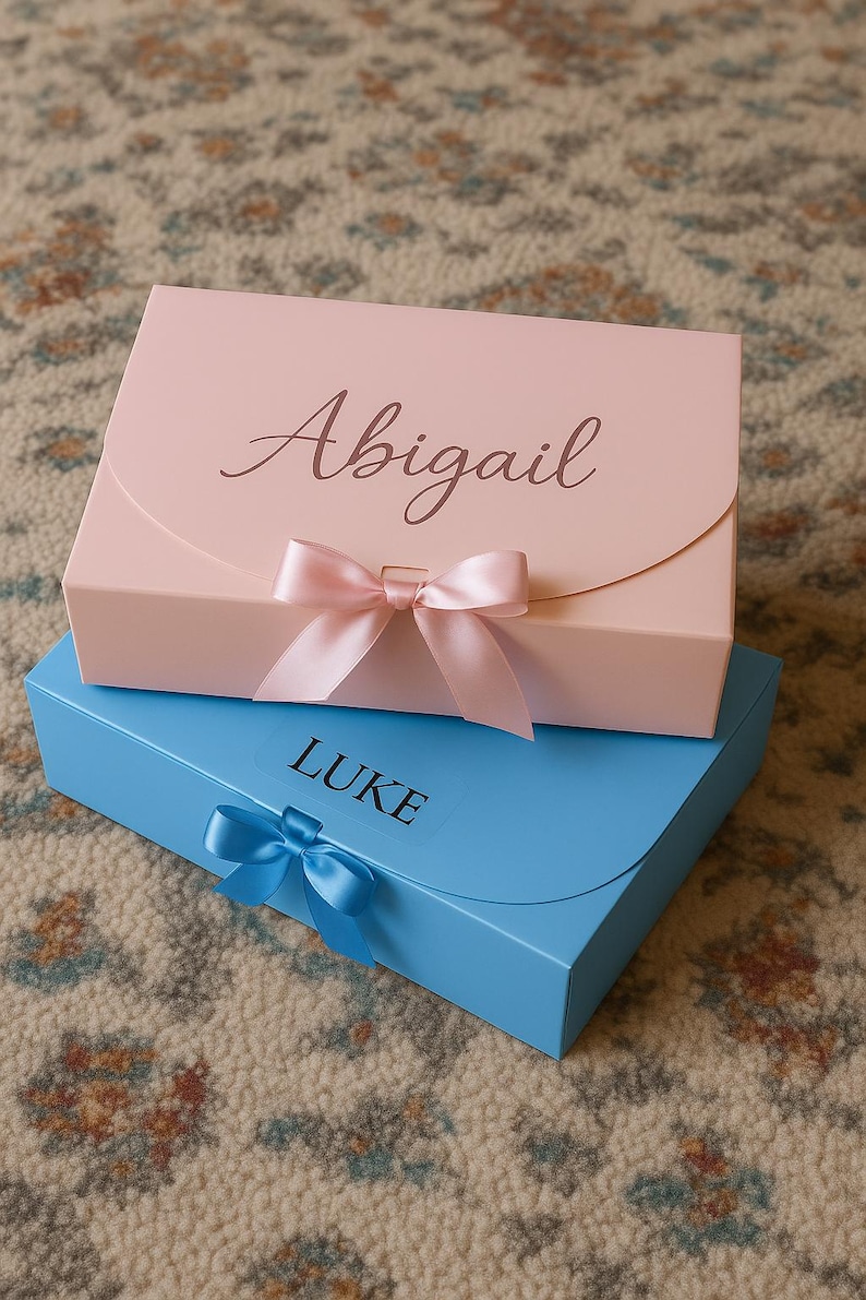 May include: Two gift boxes, one pink and one blue, stacked on top of each other. The pink box has the name "Abigail" in script, and a pink bow. The blue box has the name "LUKE" and a blue bow.