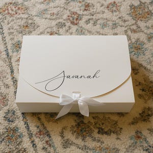 May include: A white gift box with a curved flap and a white ribbon closure. The name "Javanah" is written in elegant script on the front of the box. The box is sitting on a patterned rug.