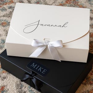 May include: Two gift boxes, one ivory and one black, are stacked. The ivory box has the name "Savannah" in cursive and a white ribbon. The black box has a "Mike" label and a black ribbon. Both boxes are closed.