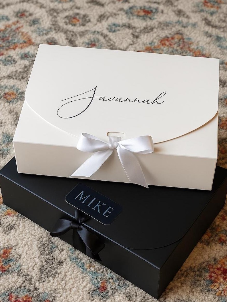 May include: Two gift boxes, one ivory and one black, are stacked. The ivory box has the name "Savannah" in cursive and a white ribbon. The black box has a "Mike" label and a black ribbon. Both boxes are closed.