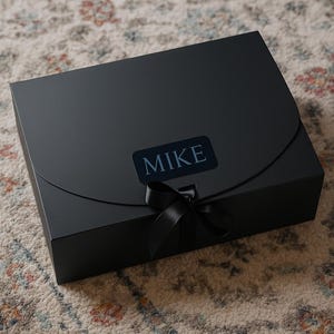 May include: A black gift box with a matching black ribbon and the name "MIKE" printed on a label. The box is rectangular with a curved flap closure. The box is sitting on a patterned rug.