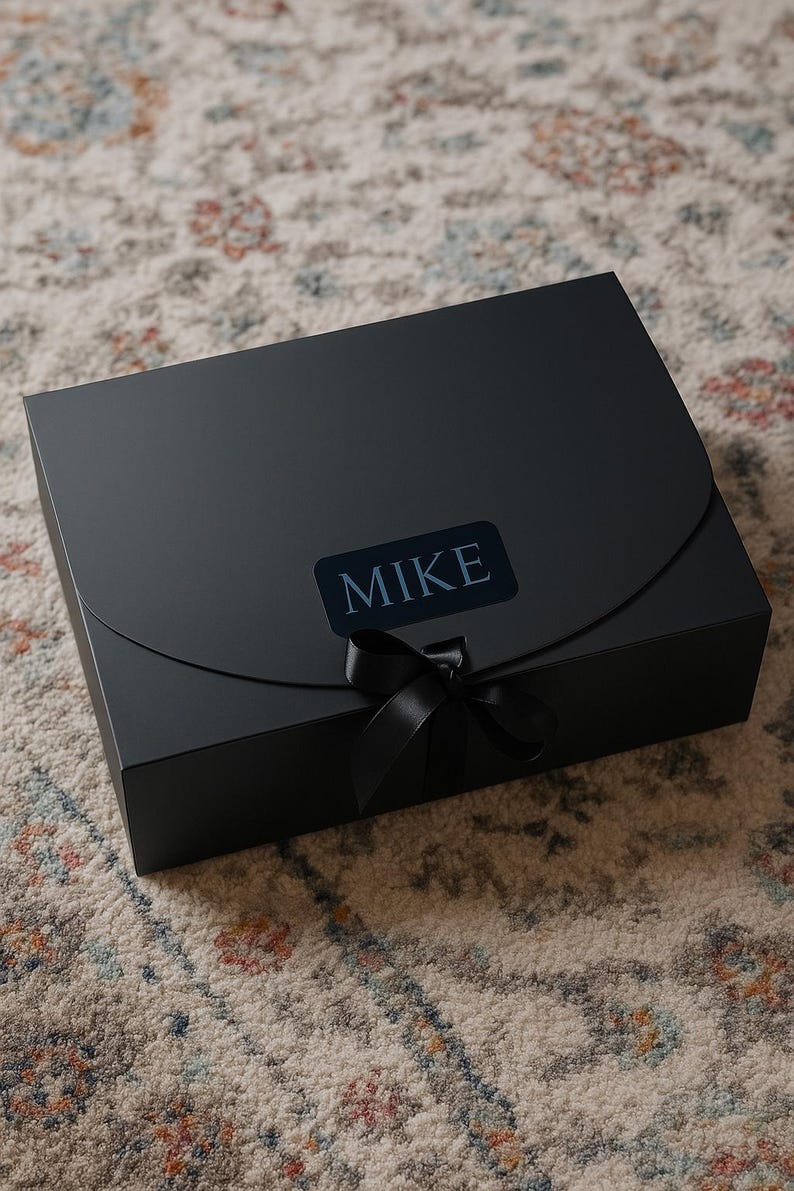 May include: A black gift box with a matching black ribbon and the name "MIKE" printed on a label. The box is rectangular with a curved flap closure. The box is sitting on a patterned rug.