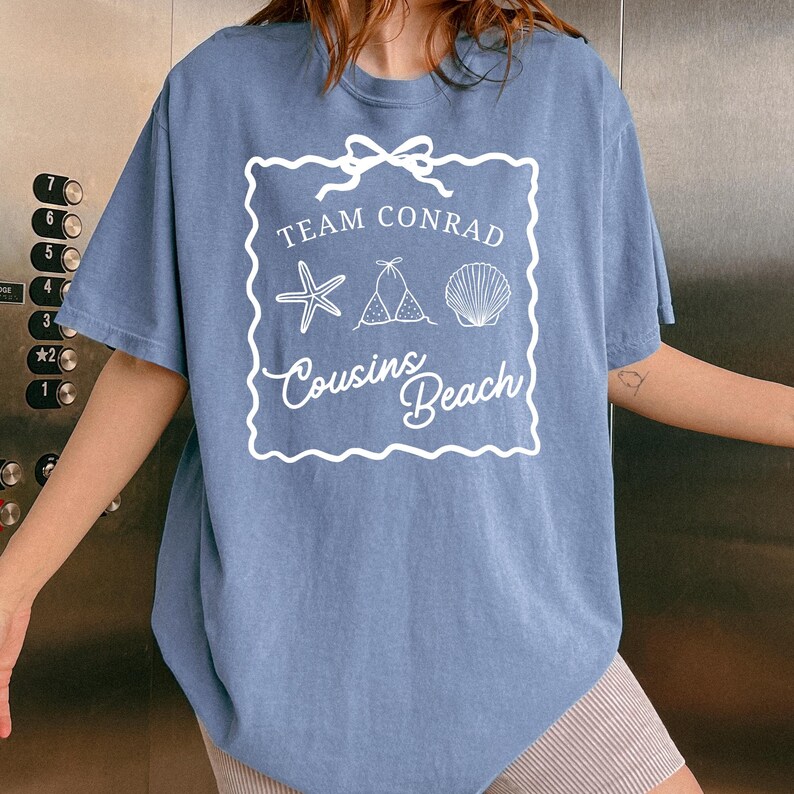 以下が含まれることがあります： 「Team Conrad Cousins Beach」の文字とヒトデ、ビキニ、貝殻の白いグラフィックがプリントされた青いTシャツ。半袖でゆったりとしたフィット感です。