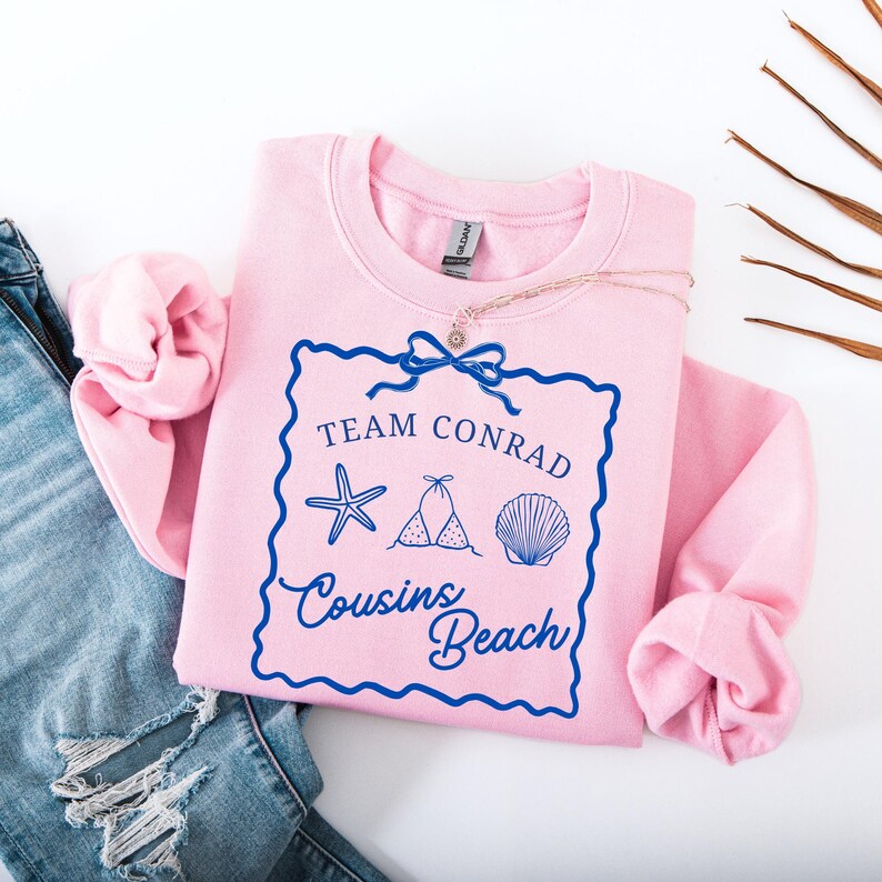 以下が含まれることがあります： 水色の文字で「Team Conrad Cousins Beach」と書かれた、ピンク色のスウェットシャツ。ヒトデ、ビキニ、貝殻のグラフィックが描かれています。繊細なネックレスがスウェットシャツの上に置かれています。スウェットシャツは、青いジーンズの隣に折りたたまれています。