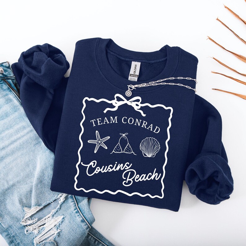 以下が含まれることがあります： 「Team Conrad Cousins Beach」の白い文字とビーチをテーマにしたグラフィックが特徴のネイビーブルーのスウェットシャツ。シルバーのネックレスがスウェットシャツの上にかけられています。スウェットシャツはブルージーンズの上に折りたたまれています。