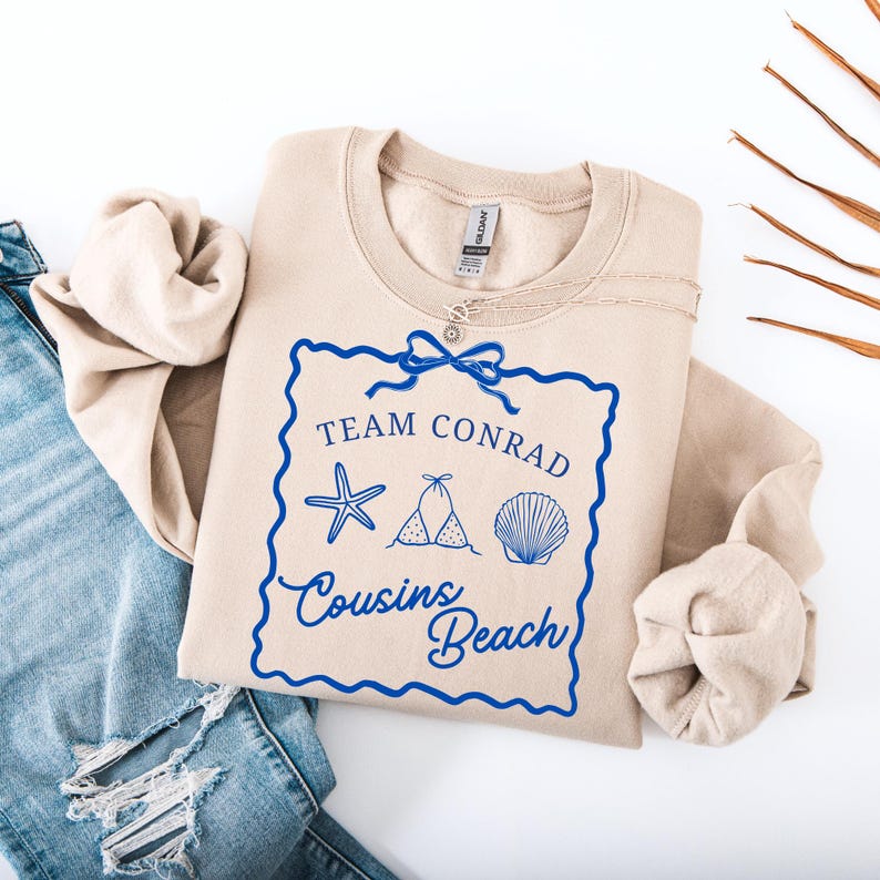 以下が含まれることがあります： 「Team Conrad Cousins Beach」の文字とヒトデ、ビキニ、貝殻のイラストが描かれた青いグラフィックのベージュのスウェットシャツ。スウェットシャツは、青いジーンズの上に折りたたまれています。