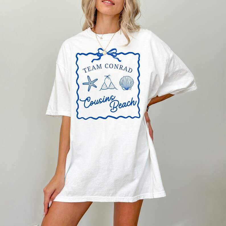 以下が含まれることがあります： 白いTシャツ。青いグラフィックには「Team Conrad」の文字、ヒトデ、ビキニ、貝殻、そして下に「Cousins Beach」と書かれています。Tシャツは半袖でゆったりとしたフィット感です。