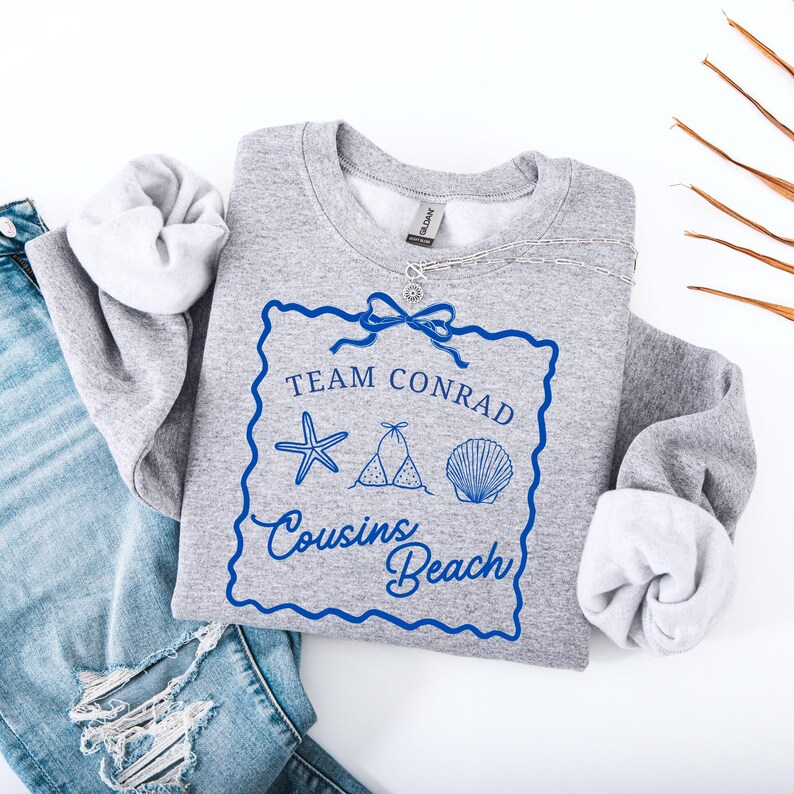 以下が含まれることがあります： 「Team Conrad Cousins Beach」の青い文字とビーチをテーマにしたグラフィックが特徴のグレーのスウェットシャツ。スウェットシャツは折りたたまれており、袖が見えています。シルバーのネックレスが上にかけられています。デザインにはヒトデ、ビキニ、貝殻が含まれています。