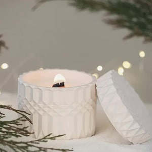 Op de afbeelding: Een brandende witte kaars in een geometrisch patroon pot met bijpassende deksel. De kaars heeft een houten lont en is omgeven door groen en zachte, onscherpe lichten, waardoor een gezellige, feestelijke sfeer ontstaat. De kaars staat op een wit oppervlak.