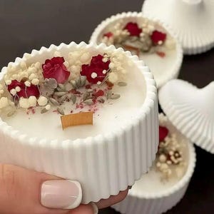 Op de afbeelding: Een witte, ronde kaars met een gegroefde rand, versierd met gedroogde rode rozen en andere botanische elementen. De kaars heeft een houten lont. Andere kaarsen en deksels staan op de achtergrond.