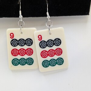 May include: A pair of ivory-colored earrings featuring a design reminiscent of a Mahjong tile. Each earring displays the number "9" in red, along with three rows of floral symbols in black, red, and green. The earrings hang from silver-colored hooks.