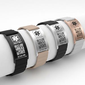 Könnte beinhalten: Eine Auswahl an medizinischen Armbändern in Schwarz, Beige, Silber und Roségold. Jedes Armband hat eine Metallplatte mit einem medizinischen Symbol, einem QR-Code und dem Text "HEALTH INFO" und "SCAN ME". Die Armbänder werden auf einer weißen zylindrischen Oberfläche präsentiert.