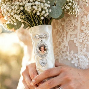 Puede incluir: Un dije de foto ovalado en tono plateado sujeto a una cinta de satén blanca envuelta alrededor de un ramo de flores. El dije presenta el retrato de una mujer y un colgante en forma de corazón con el texto grabado, Mom, I love you are always by my side.