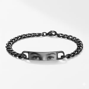 Puede incluir: Pulsera de cadena negra con una placa rectangular que muestra una imagen en escala de grises de unos ojos. La pulsera tiene un cierre de mosquetón y un diseño de cadena de bordillo. La joya se muestra sobre un fondo blanco.