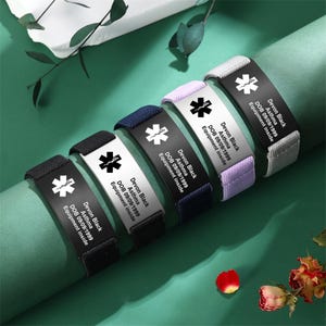 May include: A collection of medical alert bracelets in various colors, including black, navy, white, lavender, and gray. Each bracelet features a metal plate with a medical symbol and the text "Devon Black, Asthma, DOB 09/09/1999, Equipment inside."