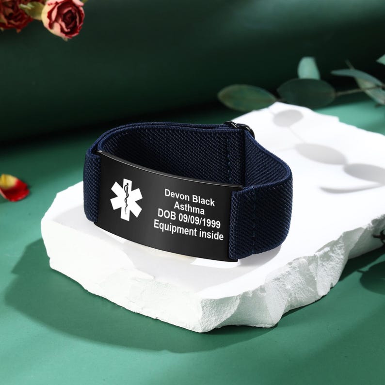 May include: A navy blue medical alert bracelet with a black plate. The plate displays a white medical symbol and text: Devon Black, Asthma, DOB 09/09/1999, Equipment inside. The bracelet is resting on a white, textured surface.
