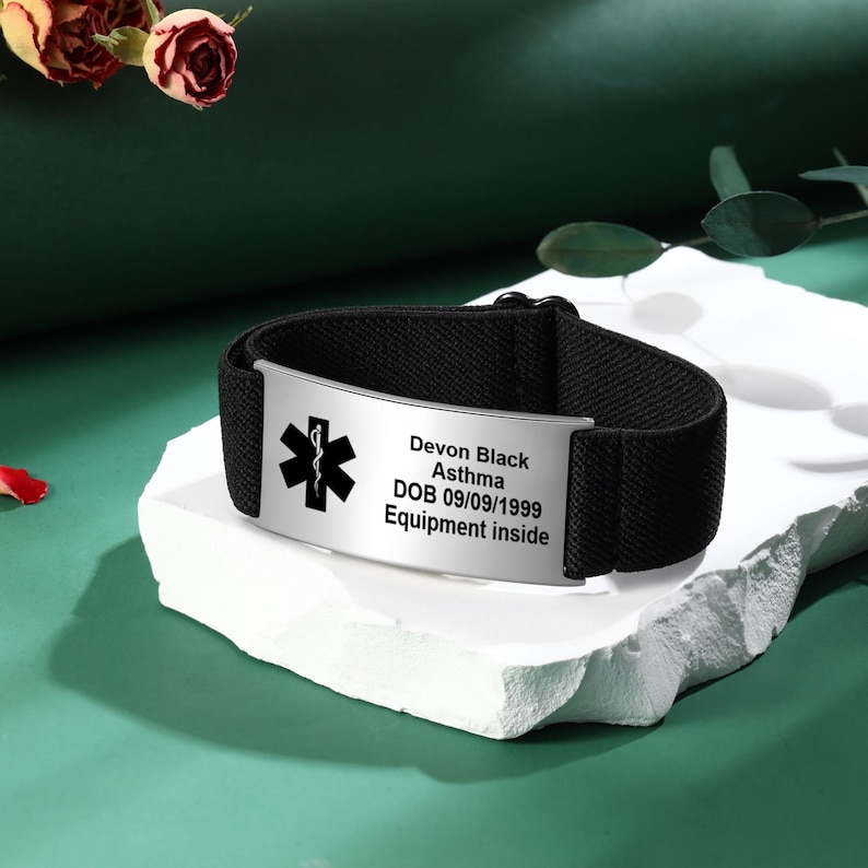 May include: Black medical alert bracelet with a silver plate. The plate has a medical symbol and the text: "Devon Black, Asthma, DOB 09/09/1999, Equipment inside." The bracelet is on a white surface against a green background.