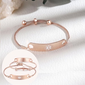Könnte beinhalten: Roségoldenes medizinischer Notfallarmband mit gebogener ID-Platte und weißem medizinischem Symbol. Das verstellbare Armband hat eine feine Kette und kleine Kugelakzente. Eine kleinere Version des Armbands ist ebenfalls abgebildet.