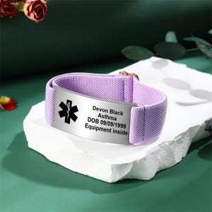 May include: A lavender medical alert bracelet with a silver plate. The plate has a black medical symbol and the text: Devon Black, Asthma, DOB 09/09/1999, Equipment inside. The band is made of a soft, woven material.