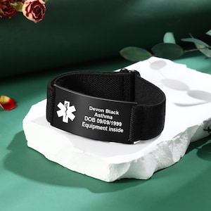 May include: Black medical alert bracelet with a white medical symbol and the text "Devon Black, Asthma, DOB 09/09/1999, Equipment inside." The bracelet is on a white surface with a green background.