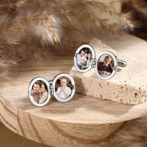 Op de afbeelding: Twee zilverkleurige, ovale manchetknopen met scharnierende frames. Elke manchetknop bevat twee kleine, ronde foto's. De ene manchetknop toont een koppel en een individu, terwijl de andere een familiegroep en een individu laat zien.