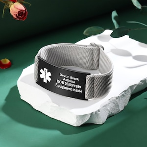 May include: A gray medical alert bracelet with a black plate. The plate has a white medical symbol and the text: Devon Black, Asthma, DOB 09/09/1999, Equipment inside. The bracelet is on a white surface against a green background.