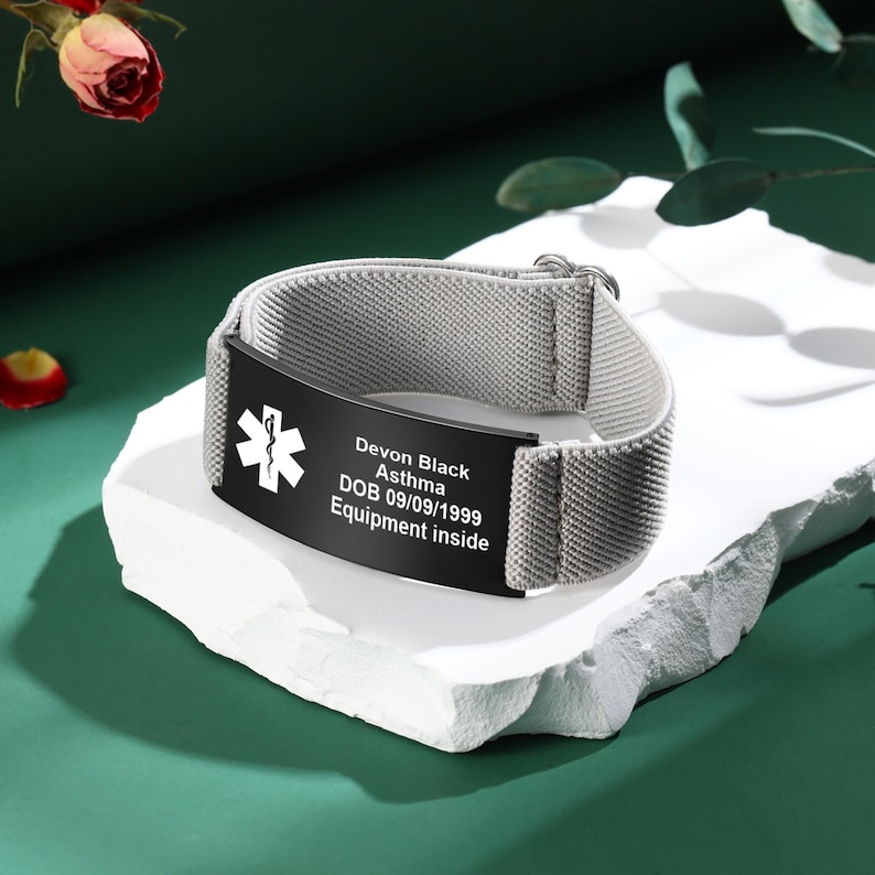 May include: A gray medical alert bracelet with a black plate. The plate has a white medical symbol and the text: Devon Black, Asthma, DOB 09/09/1999, Equipment inside. The bracelet is on a white surface against a green background.