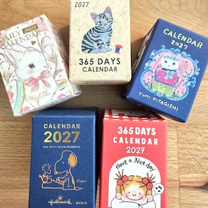 May include: Five 2027 daily calendars featuring various illustrations: a rabbit, a tabby cat, a character in a blue frame, Snoopy typing, and a girl with the text Have a Nice day. The boxes are rectangular with colorful graphic designs.
