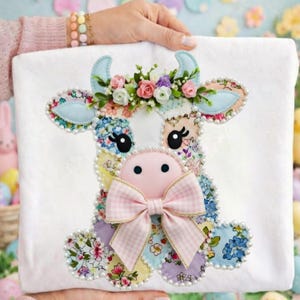Peut inclure: Carré de tissu blanc avec un motif de vache en patchwork. La vache a des patchs à motifs floraux, des oreilles bleues et un nez et un nœud roses. Une couronne florale et des perles complètent le design. La vache est tenue par une personne.