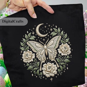 May include: Black decorative pillow with an embroidered design featuring a moth, moon, stars, and floral elements. The moth is cream-colored with green accents, surrounded by white flowers and green leaves. The text "DigitalCrafts" is in the upper left corner.