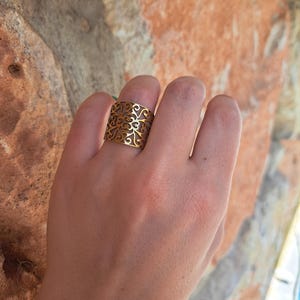 Può includere: Un anello color oro con un design traforato con motivi a vortice. L'anello è largo, coprendo una parte significativa del dito. Lo sfondo è una superficie strutturata marrone-rossastra.