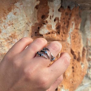 Könnte beinhalten: Ein silberfarbener Krallenring mit blauen Akzenten. Der Ring hat drei krallenartige Zacken, die von der Ringschiene ausgehen, mit kleinen blauen Details an der Basis jeder Kralle. Der Ring wird an einem Finger getragen, vor einem strukturierten, hellbraunen Hintergrund.