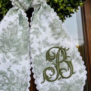 Puede incluir: Una bufanda de tela blanca con un patrón toile de jouy verde salvia y un borde de encaje blanco festoneado. La bufanda está bordada con una letra B grande de color verde oliva oscuro en una fuente cursiva decorativa y con remolinos.