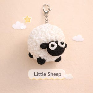 May include: A white and black crochet sheep keyring with a silver clasp. The sheep has a fluffy body, black face, and black feet. The text "Little Sheep" is below the sheep, with cloud and star decorations.
