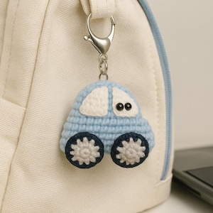 Może przedstawiać: Jasnoniebieski brelok do kluczy w kształcie szydełkowanego samochodu z białymi akcentami i ciemnoniebieskimi kołami. Samochód ma czarne guziczkowe oczy i jest przymocowany do srebrnego metalowego zapięcia. Brelok wisi na białej torbie.