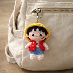 Puede incluir: Llavero de ganchillo con un personaje de pelo negro, sombrero amarillo, chaleco rojo, pantalones cortos azules y zapatos blancos. El llavero está sujeto a un cierre plateado y cuelga frente a una mochila beige.