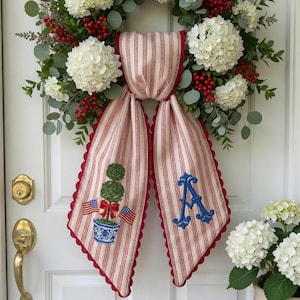 Pode incluir: Uma guirlanda decorativa com flores de hortênsia brancas, folhas de eucalipto verdes e bagas vermelhas. Uma fita de tecido creme com listras verticais vermelhas e bordas recortadas vermelhas exibe uma planta topiária bordada e uma letra A azul.
