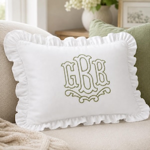 May include: A rectangular white throw pillow featuring a ruffled border and a centered olive green embroidered monogram with the letters G, B, and B in a decorative script font. The pillow has a smooth fabric texture and a classic rectangular shape.