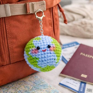 Puede incluir: Un llavero de la Tierra tejido a ganchillo con un cierre plateado y una cadena. El globo terráqueo presenta océanos azules, continentes verdes, mejillas rosas y una boca roja sonriente. El llavero está junto a una mochila y documentos de viaje.