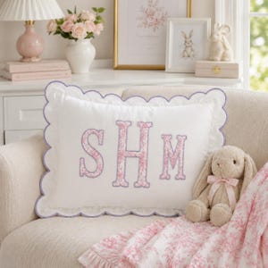 May include: A rectangular white throw pillow with a scalloped edge trimmed in lavender thread. The centre features the embroidered letters SHM in a pink floral pattern with lavender outlines. A plush tan rabbit toy sits beside the pillow.