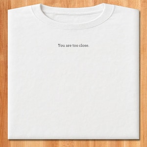 May include: White t-shirt with the text "You are too close." printed in black. The shirt has a crew neck and is folded neatly on a wooden surface. The text is centered on the front of the shirt.