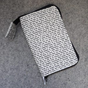 May include: A rectangular zippered wallet featuring a white fabric exterior printed with black typewriter-style text. The wallet includes a matching fabric wrist strap attached to the side, with black piping along the edges and a black zipper closure.