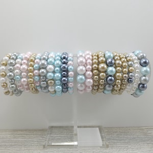 May include: A collection of pearl bracelets in various pastel colors, including pink, blue, and gray. The bracelets are stacked on a clear display stand.