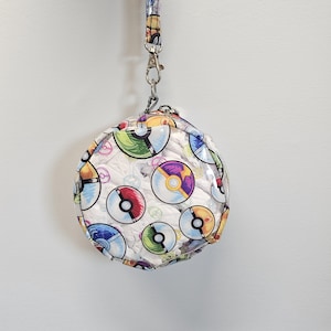 May include: A round, clear pouch with a colorful pattern of various Poke Balls. The pouch is attached to a lanyard with a silver clasp. The background is a plain white wall.