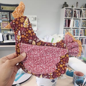 May include: A fabric chicken-shaped ornament featuring a dark maroon base with a repeating pattern of yellow flowers and small gnomes. It has a pink wing with a white botanical print, an orange comb, and a matching orange tail with a white leaf pattern.
